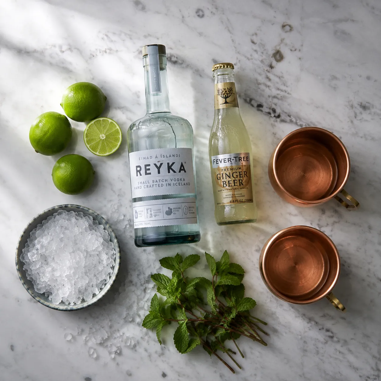 Moscow mule recipe ingredients flatlay with vodka, ginger beer, limes, and copper mugs