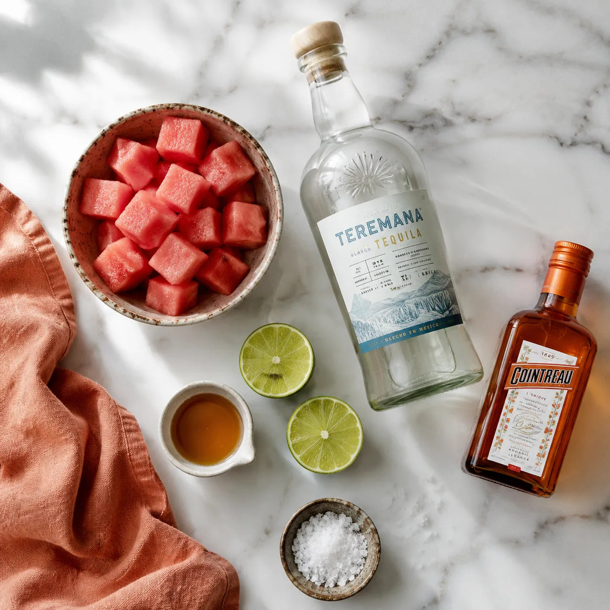 Watermelon margarita ingredients flatlay with tequila, lime, watermelon, and orange liqueur